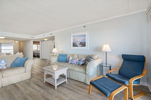 A spacious living area with a beige sofa, a blue armchair, and a wicker coffee table. A dining area is visible in the background with a kitchen counter and chairs.