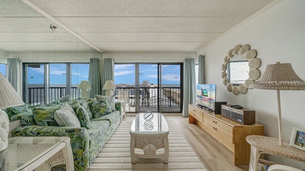 A spacious living room with a large sofa, a coffee table, and a TV. There is a balcony with a view of the city.