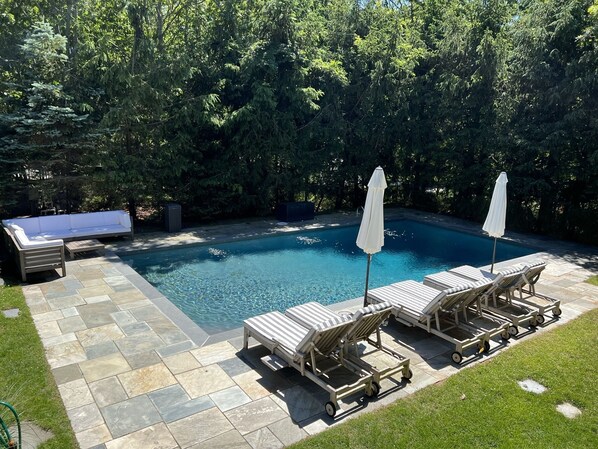 A swimming pool with lounge chairs and a white umbrella, surrounded by a stone patio and lush greenery.