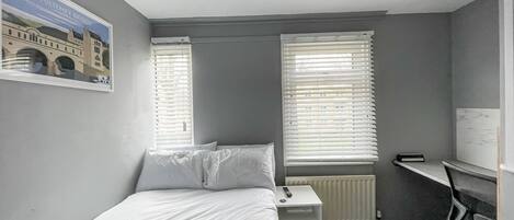 A hotel room with a bed, a desk, and two windows with blinds.