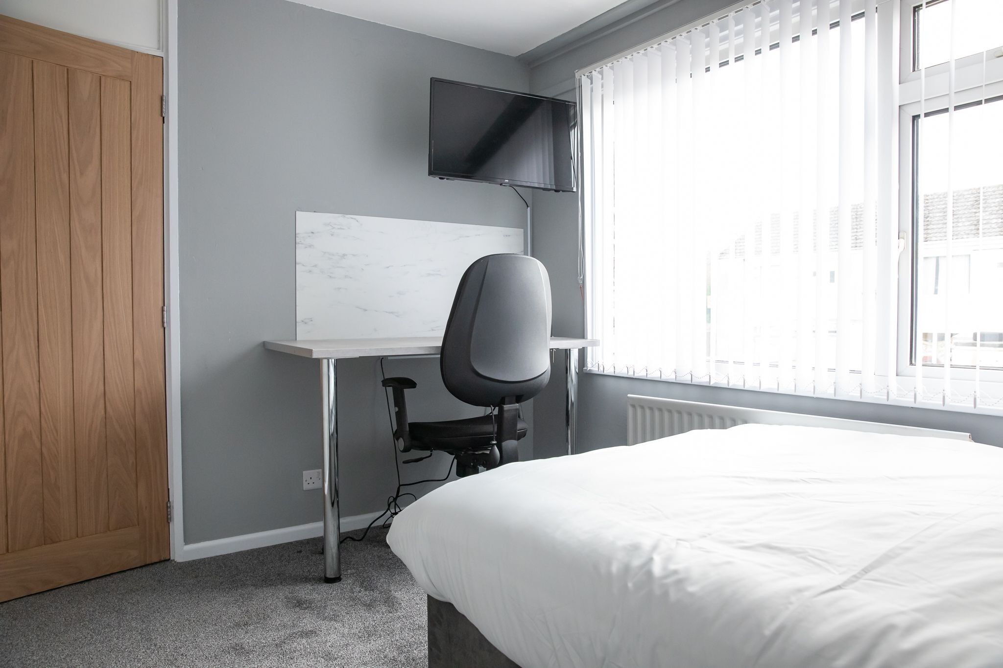 Un dormitorio con cama, escritorio con silla, televisor montado en la pared y ventana con persianas.