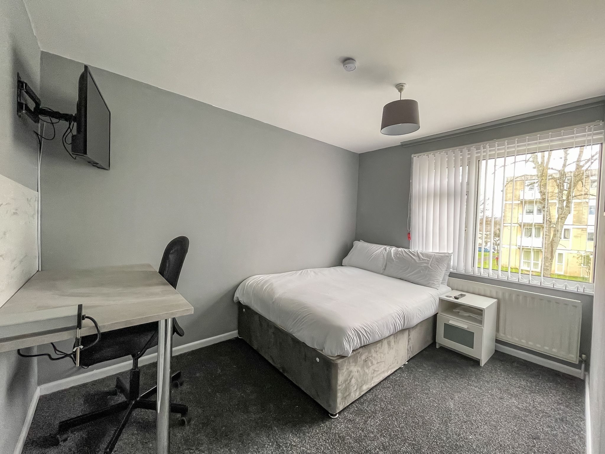 A bedroom with a bed, a desk, and a chair. There is a window with blinds and a view of buildings outside.