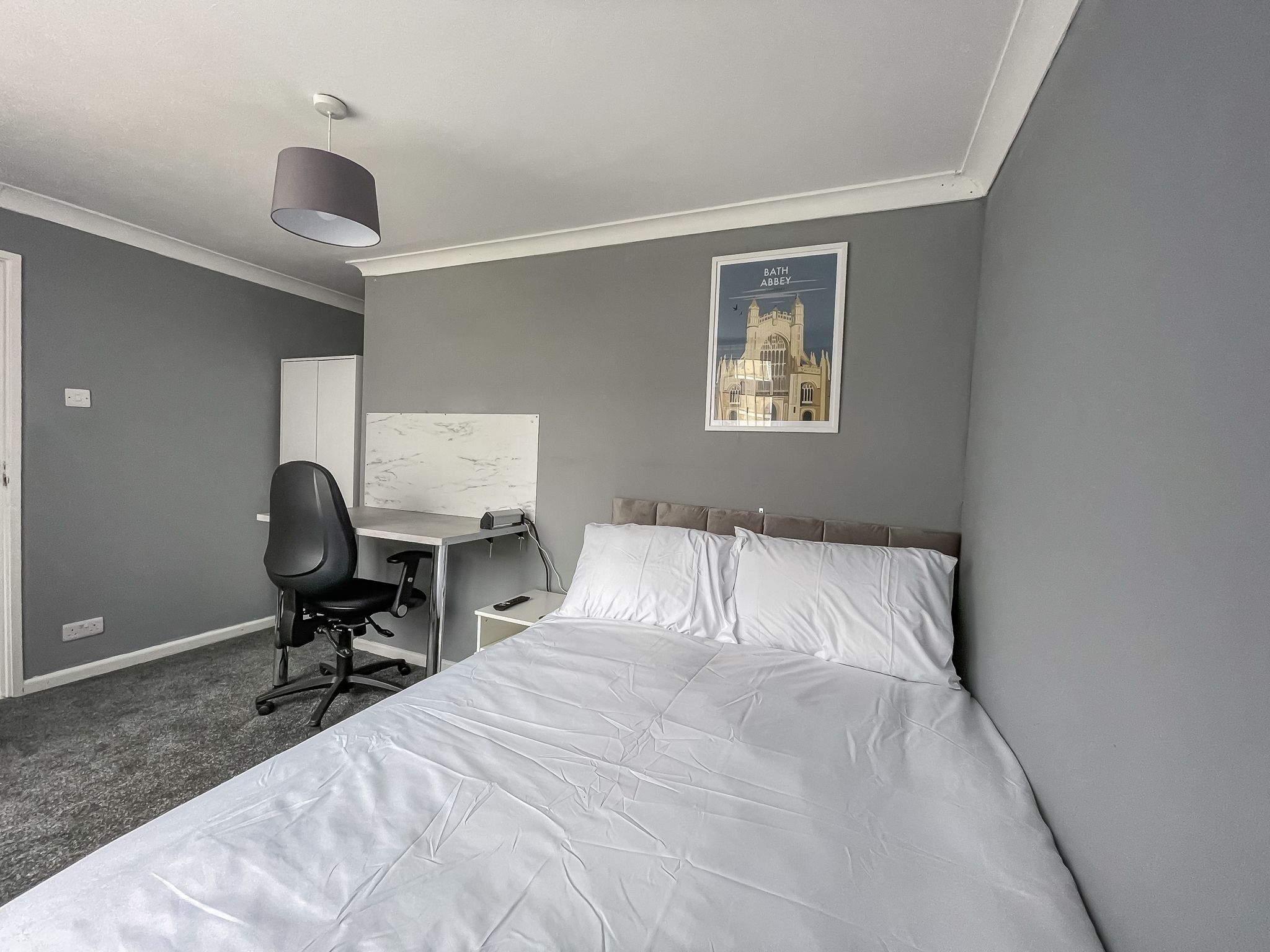 A hotel room with a bed, a desk, a chair, a lamp, and a poster of a cathedral.