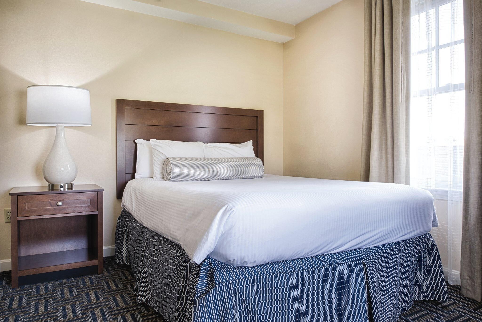 A hotel room with a large bed, a nightstand with a lamp, and a window with curtains.