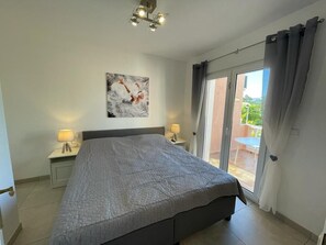 A bedroom with a bed, a nightstand, a lamp, a painting, and a sliding glass door to an outdoor area.