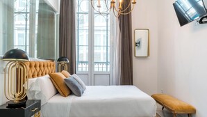 A modern bedroom with a tufted headboard, a large window with curtains, a black and gold lamp, a bench, and a flat-screen TV mounted on the wall.