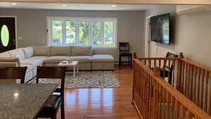 Ein Wohnzimmer mit einer Eckcouch, einem Esstisch mit Stühlen, einem gemusterten Teppich und einer Holztreppe.