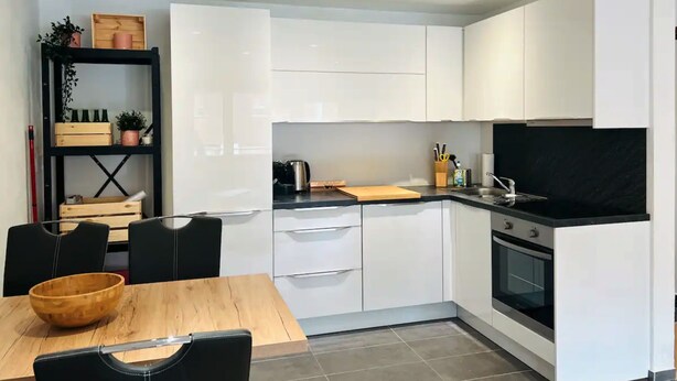 Une cuisine moderne avec des meubles blancs, un plan de travail noir et une table à manger en bois, accompagnée de chaises.