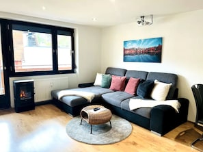 A modern living room with a grey sofa, a small round ottoman, a fireplace, and a large window with a view of a building.