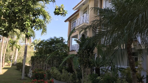 Un sendero conduce a un edificio con balcón, rodeado de exuberante follaje y palmeras.