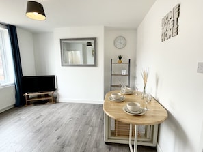 A modern living room with a wooden dining table set for two, a flat-screen TV, a mirror, a clock, and a bookshelf.