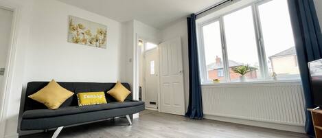 A modern living room with a grey sofa, two yellow cushions, a ceiling light, a large window with curtains, and a radiator.