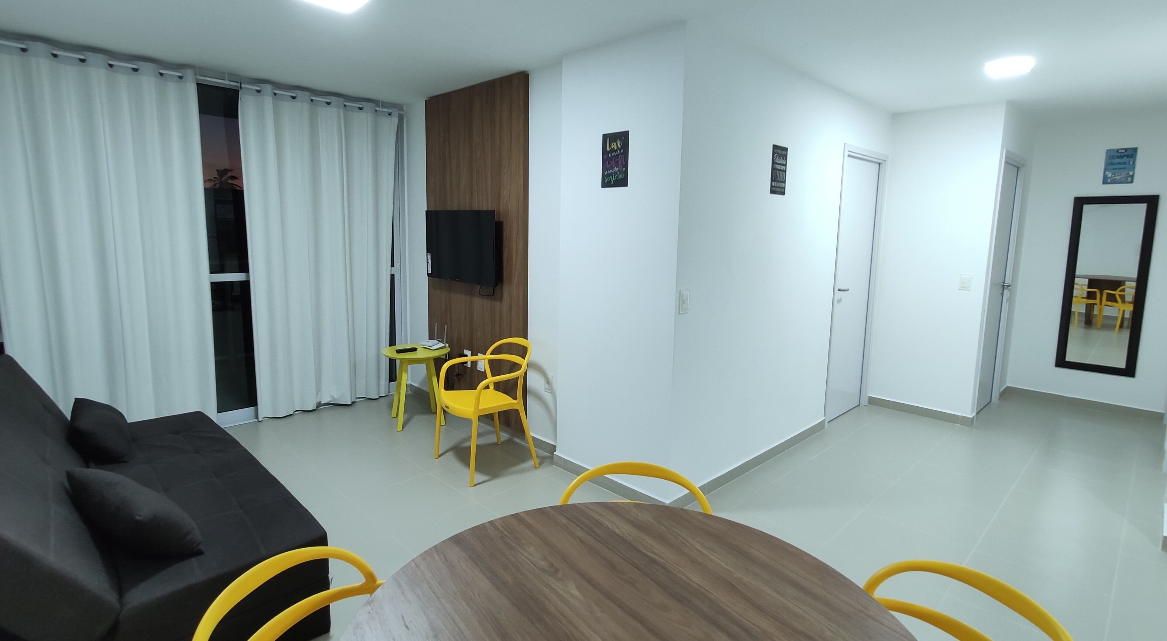A modern living room with a dark sofa, a wooden coffee table, and yellow chairs. There is a small table with a remote control and a book. The room has white walls, a wooden accent wall, and a large window with white curtains.