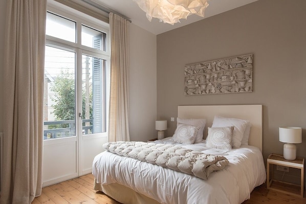 A bedroom with a large bed, two bedside lamps, a window with curtains, and a wall art piece.