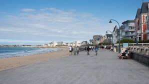 A coastal promenade with a beach, people walking, and buildings along the shore.
