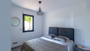 Un dormitorio moderno con una cama grande, un espejo redondo, una ventana con vistas a zonas verdes y una lámpara colgante geométrica original.