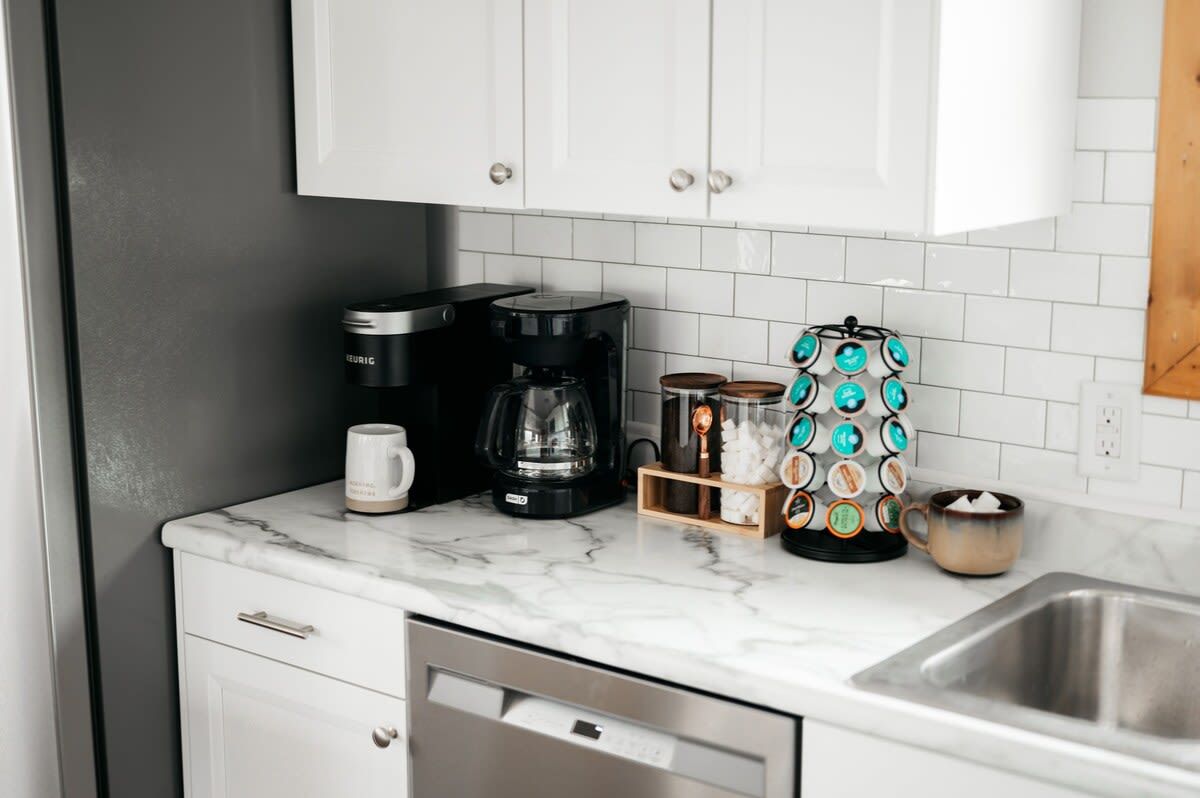 A kitchen with white cabinets, a coffee maker, a coffee pot, and a coffee machine.