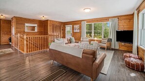 A wooden cabin interior with a brown sofa, a TV, and a staircase leading to an upper level. - Hot Tub! | Pet Friendly! | Game Room! (Traverse City)