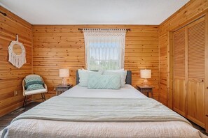 A wooden-paneled bedroom with a bed, two bedside tables, a chair, and a window with curtains. - Hot Tub! | Pet Friendly! | Game Room! (Traverse City)