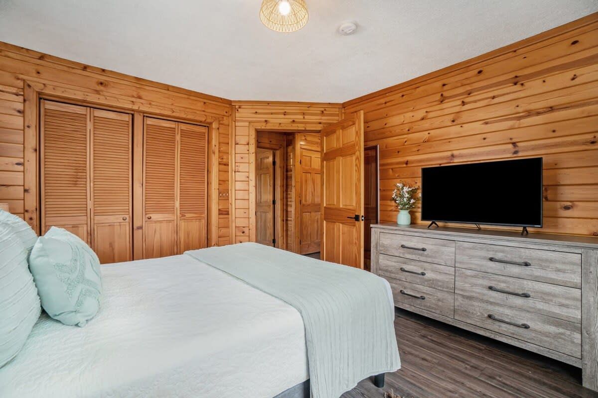A bedroom with wooden walls, a bed with pillows, a dresser, and a television.