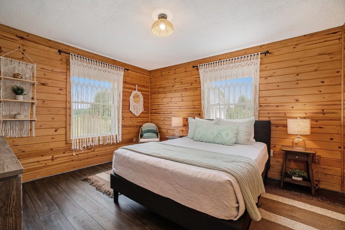 A wooden-paneled bedroom with a bed, two bedside tables, a hanging macrame wall decoration, and a window with lace curtains.