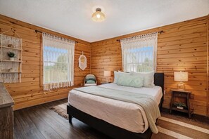 A wooden-paneled bedroom with a bed, two bedside tables, a hanging macrame decoration, and a small shelf with a plant.