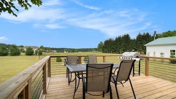 A wooden deck with a table and chairs, a grill, and a view of a green landscape.