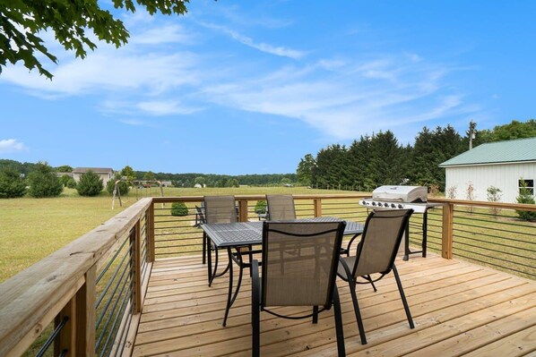 A wooden deck with a table and chairs, a grill, and a view of a green landscape.