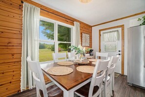 A wooden-paneled room with a dining table set for six, white chairs, a refrigerator, and a window with curtains. - Hot Tub! | Pet Friendly! | Game Room! (Traverse City)