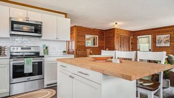 Una cucina con mobili bianchi, un'isola in legno, elettrodomestici in acciaio inossidabile e una finestra con persiane.