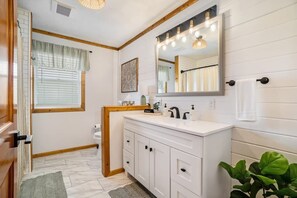 A bathroom with a white vanity, a large mirror with lights, a toilet, and a window with blinds. - Hot Tub! | Pet Friendly! | Game Room! (Traverse City)