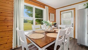 A wooden-paneled room with a dining table set for four, white chairs, a refrigerator, and large windows offering a view of greenery. - Hot Tub! | Pet Friendly! | Game Room! (Traverse City)