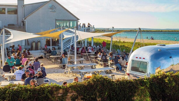 A beachside gathering with picnic tables and a large building with a dragon emblem. - Spacious Retreat With Pool, Game Room & Fireplace | Sleeps 10 | Backyard Oasis & Screened Gazebo | Family-Friendly Getaway | 24/7 Hospitality (Plymouth)