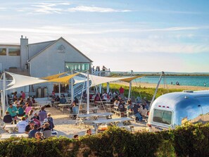 Une réunion conviviale sur la plage, avec des tables de pique-nique et un grand bâtiment orné d’un emblème de dragon.