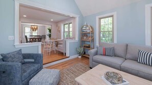 A spacious living room with a grey sofa, a blue armchair, and a wooden coffee table. There is a dining area with a table and chairs visible through an open doorway. - Spacious Retreat With Pool, Game Room & Fireplace | Sleeps 10 | Backyard Oasis & Screened Gazebo | Family-Friendly Getaway | 24/7 Hospitality (Plymouth)