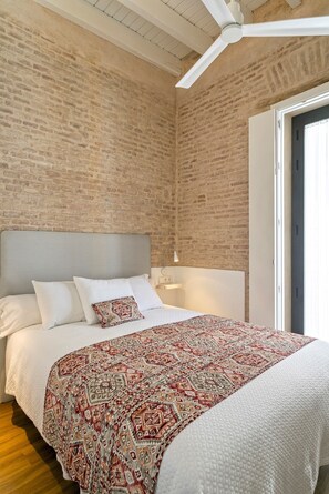 Een slaapkamer met een bakstenen muur, een bed met witte beddengoed, een geborduurde plaid, een plafondventilator en een nachtkastje met een lamp.