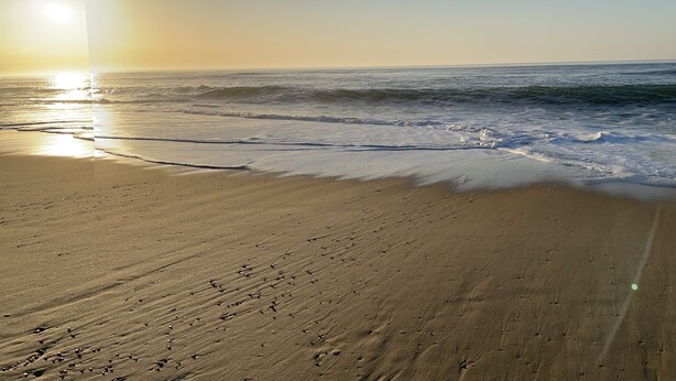 Ein Strand mit blauem Himmel und Sonnenlicht, das sich im Wasser spiegelt.