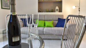 A bottle of wine, two wine glasses, and a chair on a glass table in a living room with a sofa and decorative items in the background. - Benedettini - Suites (Catania)