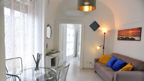A modern living room with a glass dining table, a sofa with colorful cushions, and a painting of a sunset. - Benedettini - Suites (Catania)