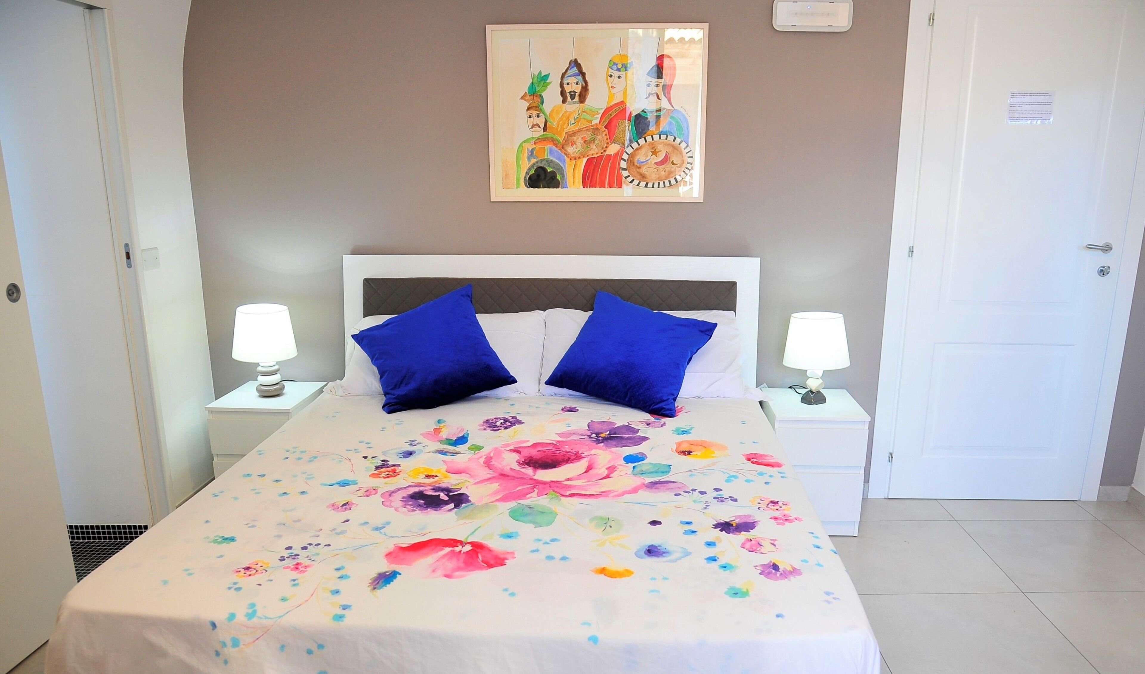 A hotel room with a bed, two blue pillows, a floral bedspread, two bedside lamps, a painting on the wall, and a door.