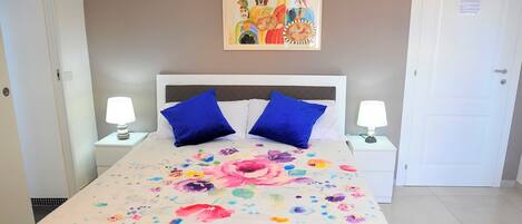A hotel room with a bed, two blue pillows, a floral bedspread, two bedside lamps, a painting on the wall, and a door.