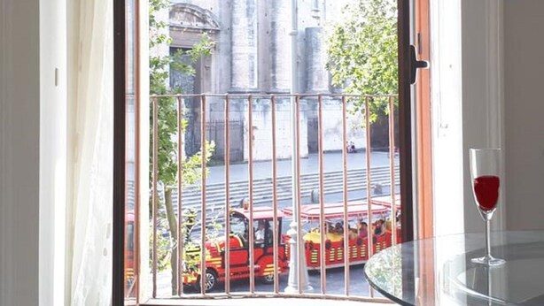 A balcony with a glass table and a wine glass, overlooking a street with a red tram. - Benedettini - Suites (Catania)