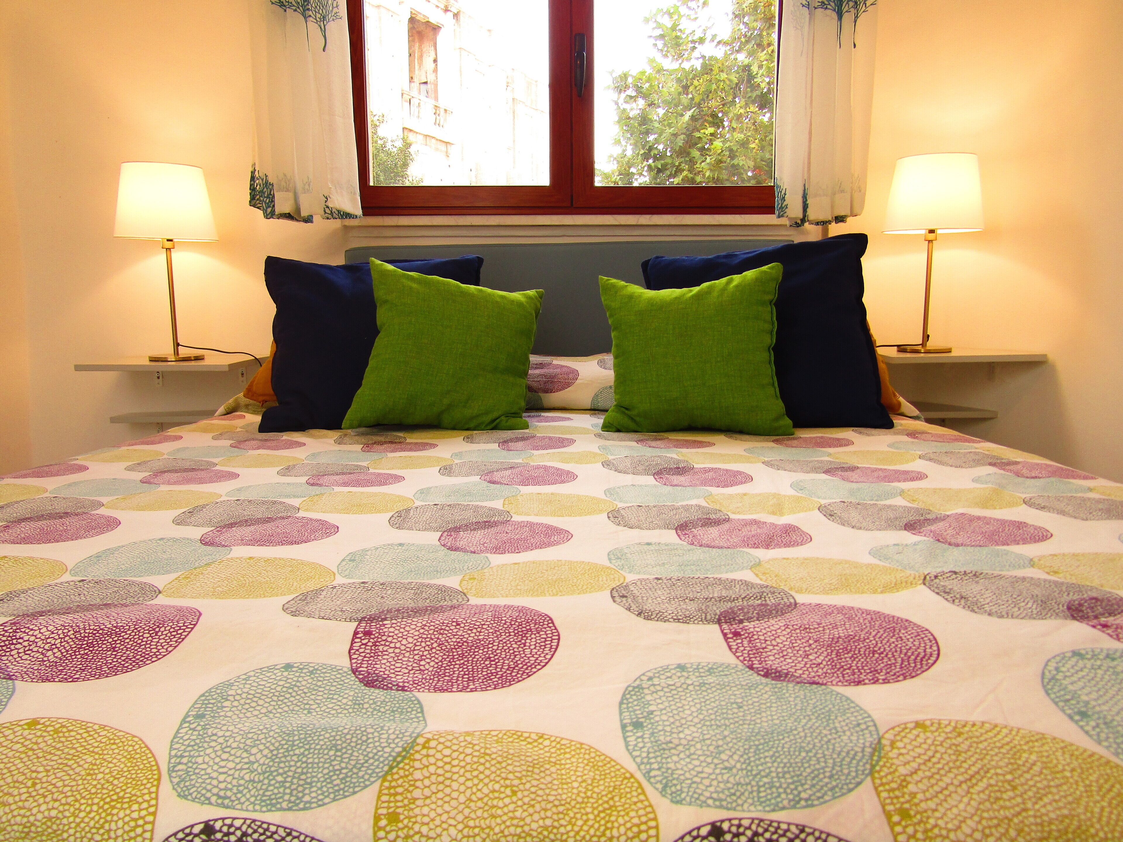 A bed with a colorful circular pattern bedspread, two green pillows, and two bedside lamps.