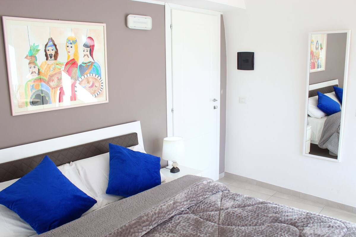 A bedroom with a bed, blue pillows, a grey quilt, a bedside lamp, a door, and a wall painting.