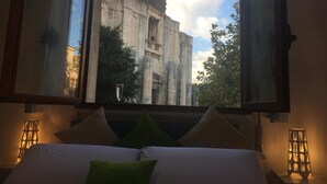 A hotel room with a bed, pillows, a window with a view of a historic building, and two wall-mounted lamps. - Benedettini - Suites (Catania)