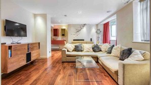 Ein modernes Wohnzimmer mit Holzboden, einem beige farbenen Sofa, einem Glastisch und einem flachbildfernseher, der an der Wand montiert ist.