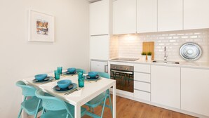 Una cocina moderna con armarios blancos, horno integrado y un lavamanos. Hay una mesa de comedor con sillas azules y platos.