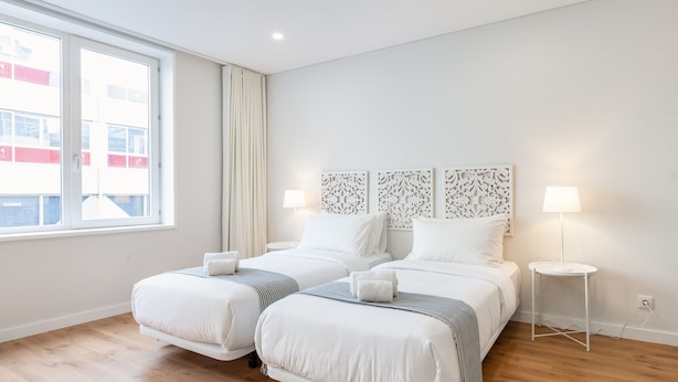 Habitación de hotel con dos camas, una ventana con cortinas, una mesita de noche pequeña con lámpara y un cabecero decorativo.