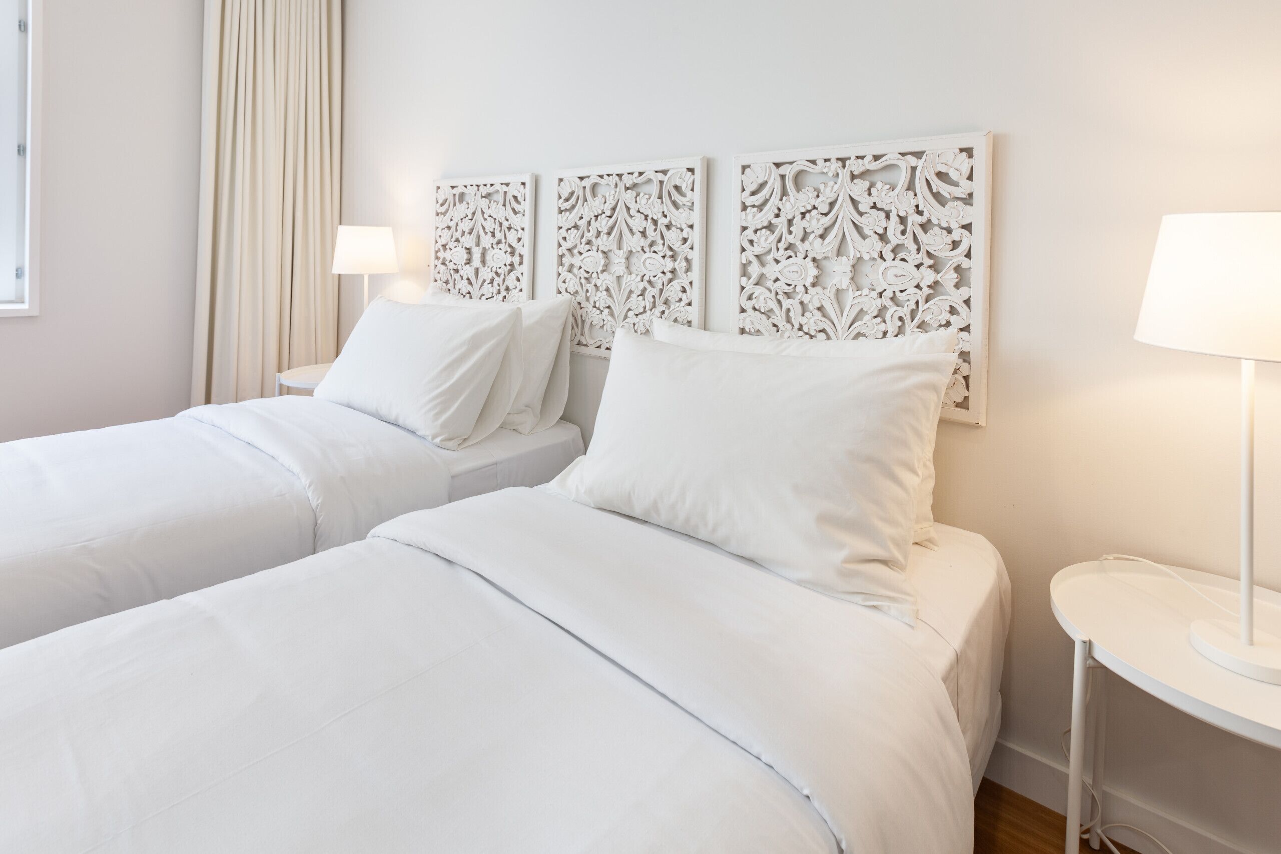 A hotel room with two beds, white bedding, a decorative headboard, a bedside table with a lamp, and a window with curtains.