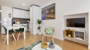 A modern kitchen with white cabinets, a dining area with a white table and green chairs, a living room with a flat-screen TV, and a cityscape painting on the wall.